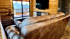 Un acogedor interior de cabaña de madera con un sofá de cuero, un televisor de pantalla plana montado en la pared y un ventanal corredizo que da a una terraza exterior.