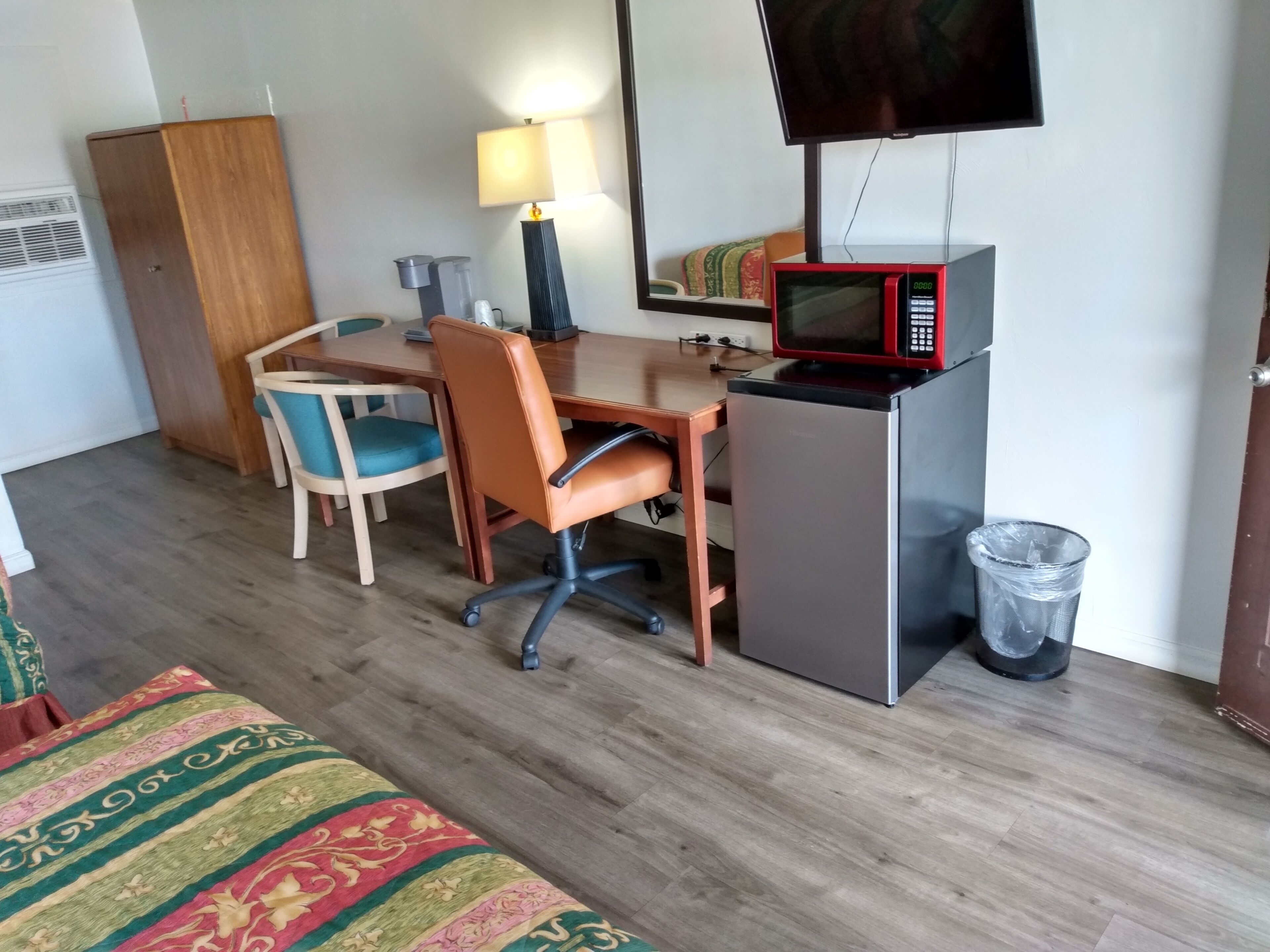 Une chambre d’hôtel avec un bureau en bois, une chaise, une télévision, un four à micro-ondes, une poubelle et une couvre-lit à motifs.