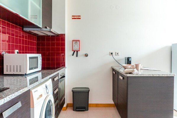 Compact kitchen with microwave, washing machine, and dishwasher, featuring a sink, countertop, and trash bin.
