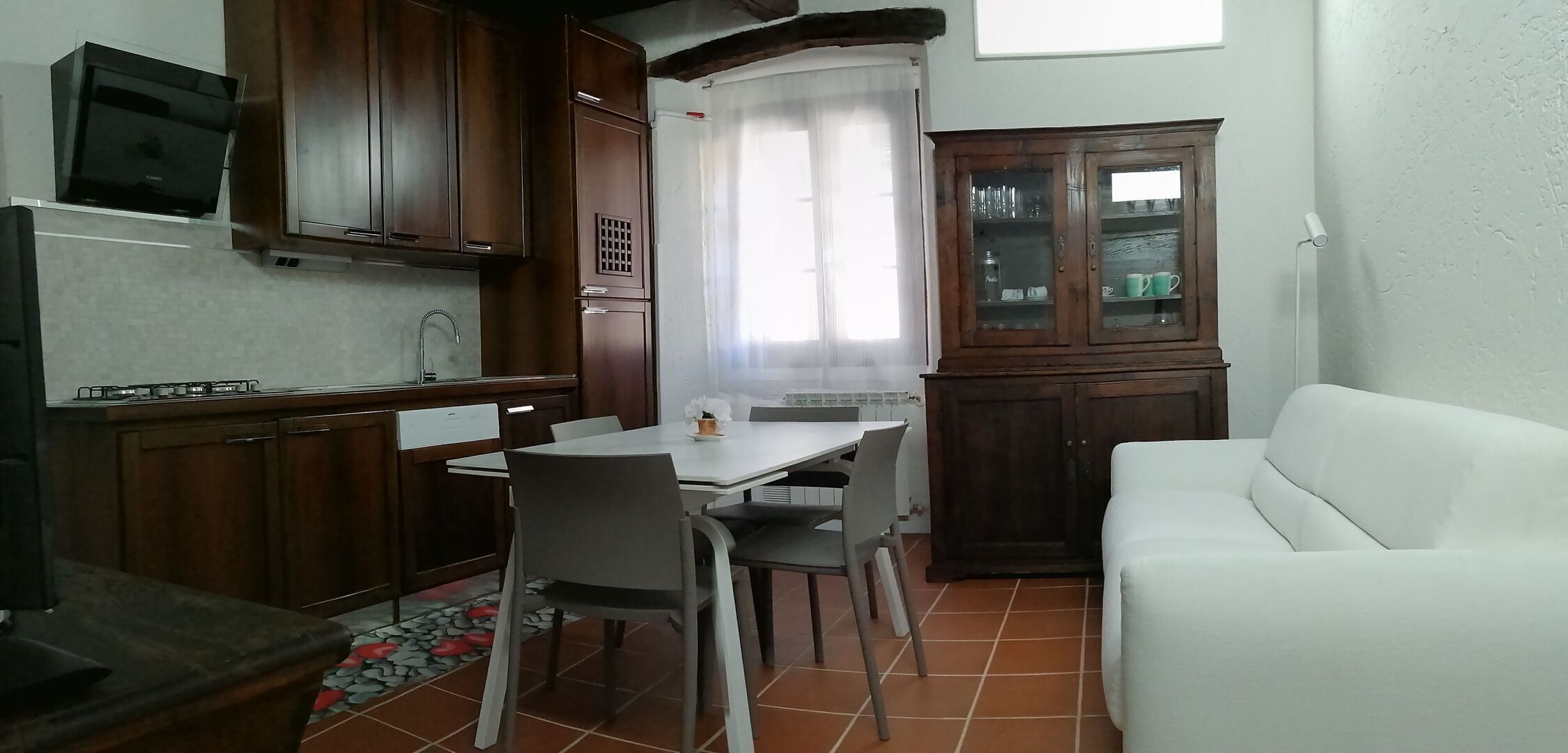 A compact kitchen with wooden cabinets, a dining table with chairs, a white sofa, and a cabinet with glass doors.