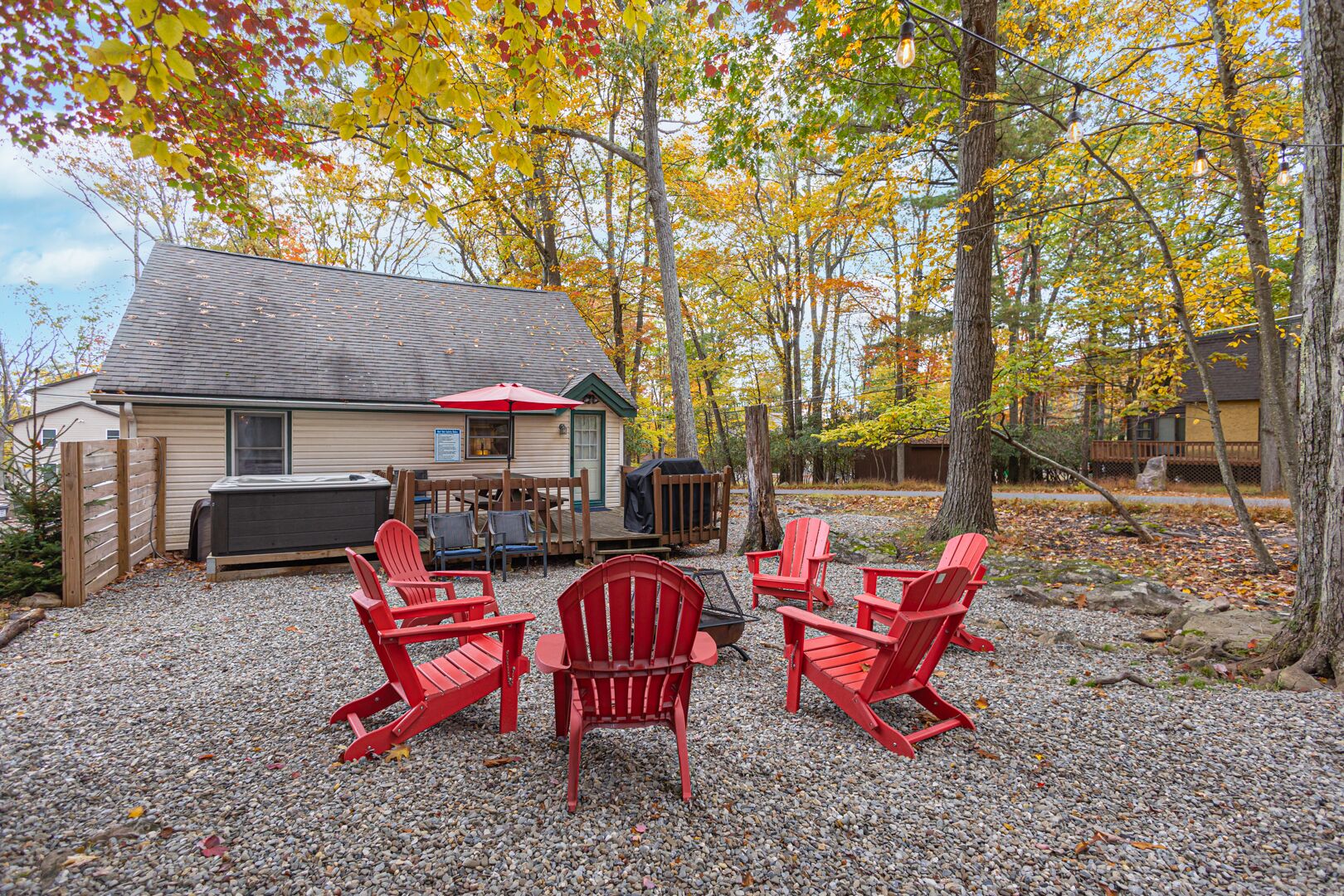 A cabin with a gravel patio, red Adirondack chairs, a red umbrella, and a hot tub.