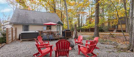A cabin with a gravel patio, red Adirondack chairs, a red umbrella, and a hot tub.