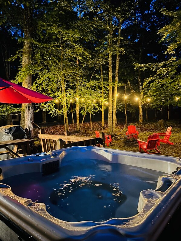 Un jacuzzi rodeado de árboles, mobiliario exterior e iluminación con luces colgantes.