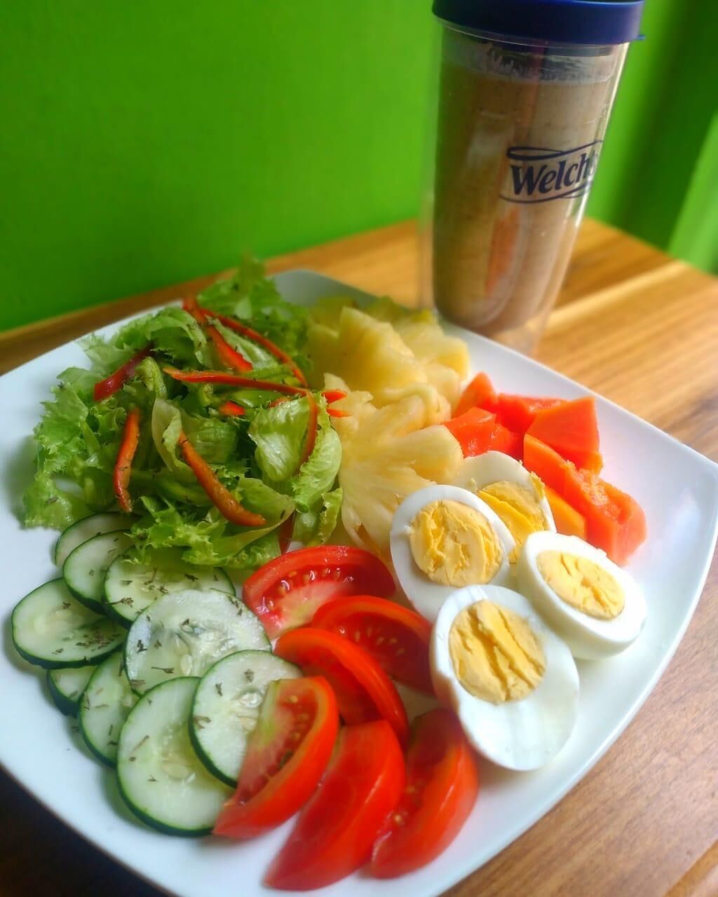 En tallerken med salat, skivede agurker, tomater, røde peberfrugter, kogte æg og en drik i et blåt glas.