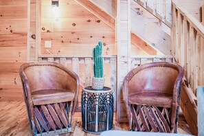 Twee leren fauteuils met een geweven onderstel, een cactus in een geweven pot, en een houten trap in een kamer met houten lambrisering.