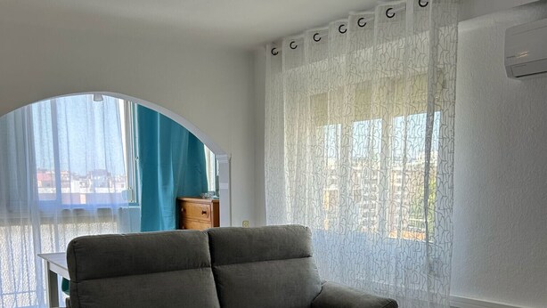 Una sala de estar con un sofá gris, una pequeña mesa de centro blanca y una ventana con cortinas traslúcidas.