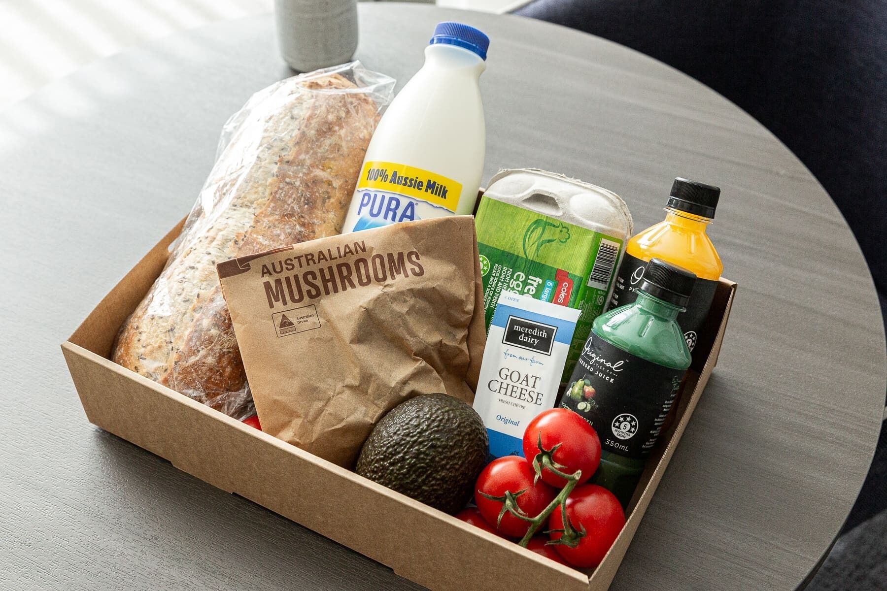 A cardboard box containing Australian mushrooms, a bottle of milk, a carton of goat cheese, a bottle of juice, and a bag of bread.