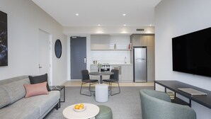 A modern living room with a sofa, ottoman, round table, and a flat-screen TV. - Quest Watergardens (Taylors Lakes)