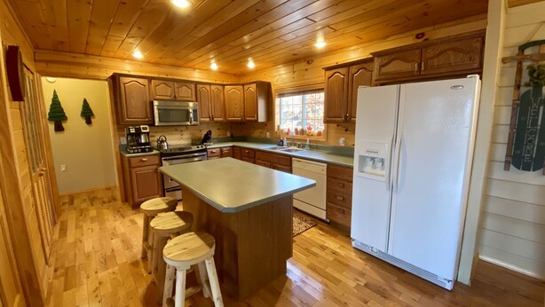 A kitchen with wooden cabinets, a central island with stools, a refrigerator, and a microwave. - ADK Lake House Vacation, Cozy 5 Bedroom & 2 Bath (Old Forge)