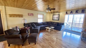 A spacious living room with a wooden ceiling and a large window overlooking a deck. - ADK Lake House Vacation, Cozy 5 Bedroom & 2 Bath (Old Forge)