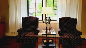 Una habitación con dos sillones estampados, una mesita con una lámpara y una planta, y un ventanal con cortinas.