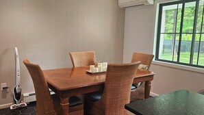 Comedor con mesa de madera, sillas de mimbre, un equipo de aire acondicionado en la pared y ventana con vista a follaje.
