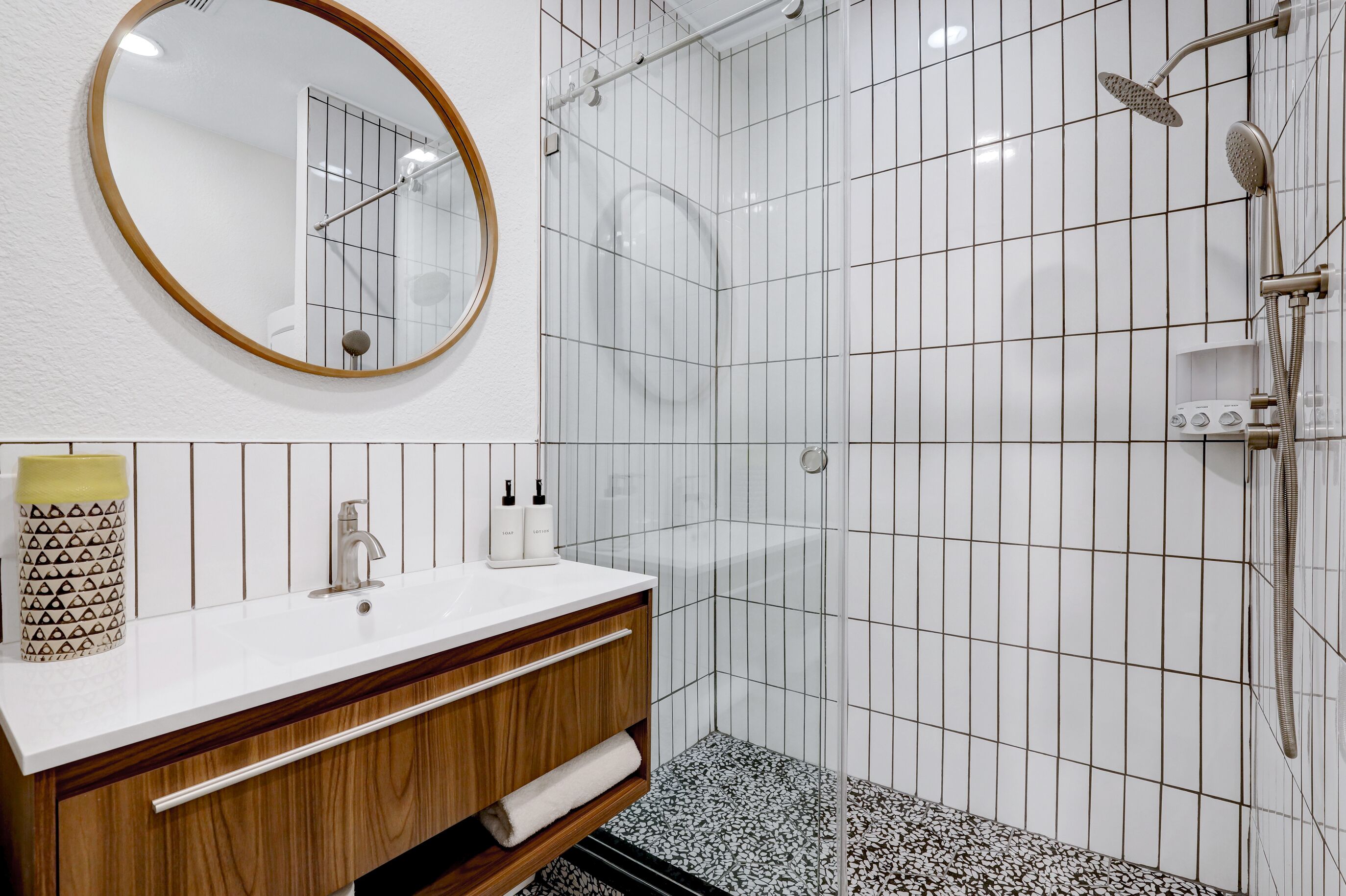 A modern bathroom with a white tiled shower, a wooden vanity with a mirror, and a white sink.