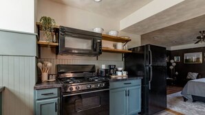 Une cuisine compacte équipée d’une cuisinière, d’un four à micro-ondes et d’un réfrigérateur. On trouve des étagères en bois au-dessus de la cuisinière et une plante sur le côté gauche. La cuisine est attenante à une chambre à coucher qui comprend un lit et une table de chevet.
