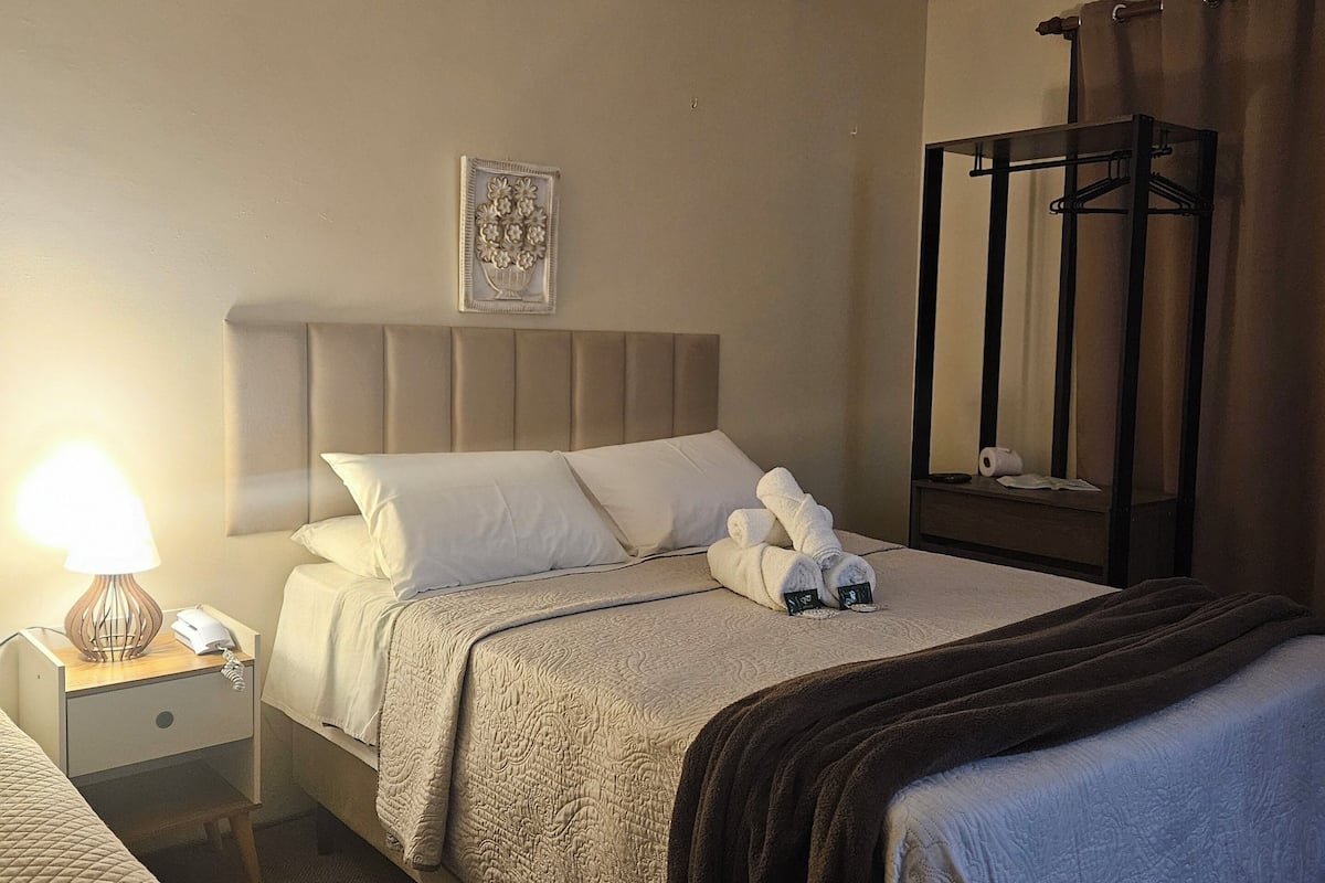 Une chambre d’hôtel avec deux lits, une table de chevet avec une lampe, un ventilateur de plafond et un tableau encadré au mur.