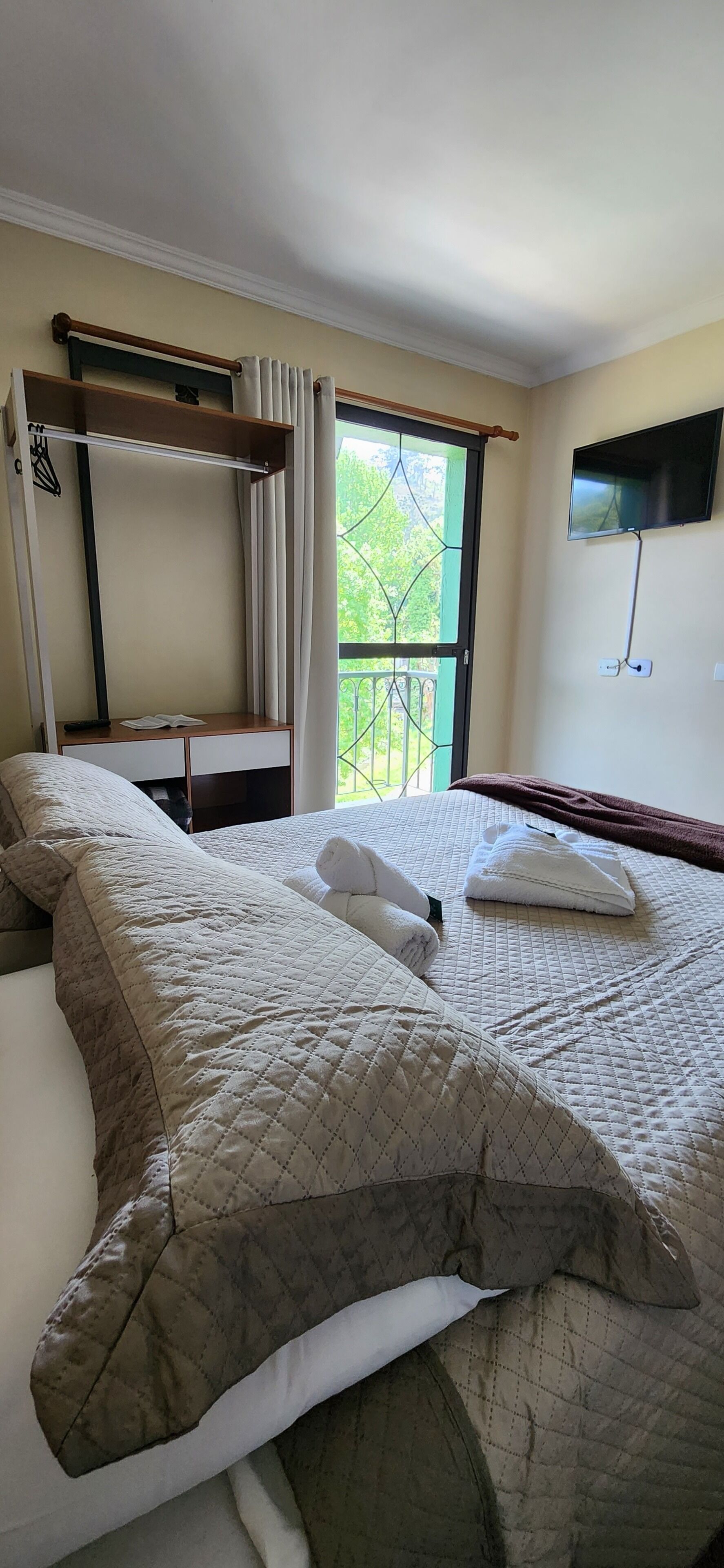 Une chambre d’hôtel avec un lit, une fenêtre avec des rideaux, un téléviseur fixé au mur et un balcon avec une balustrade.