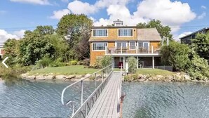 Une maison à deux étages avec un balcon, une jetée s’avançant dans l’eau et une zone herbeuse avec des arbres.
