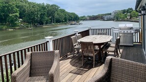 Une terrasse dotée de meubles en osier et d’un barbecue, offrant une vue sur une rivière où l’on peut apercevoir des bateaux et une végétation luxuriante.