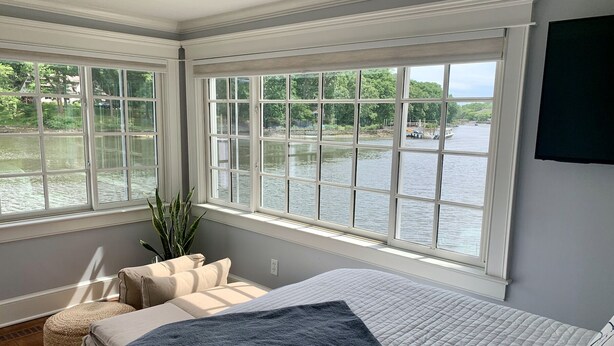 Une chambre avec une grande fenêtre donnant sur un plan d’eau, un lit recouvert d’une courtepointe matelassée, une table de chevet et une plante.