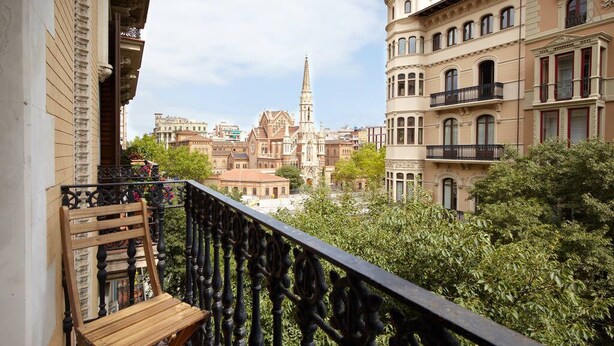 A balcony with a wooden chair, a wrought iron railing, and a view of historic buildings and a church spire. - Apartment Valencia - Two Bedroom Apartment (Barcelona)