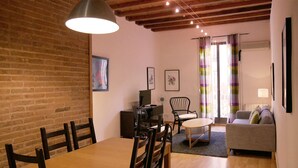 A cozy living room with a wooden dining table, chairs, a sofa, and a TV. There are framed pictures on the wall and a window with curtains. - Apartment Valencia - Two Bedroom Apartment (Barcelona)