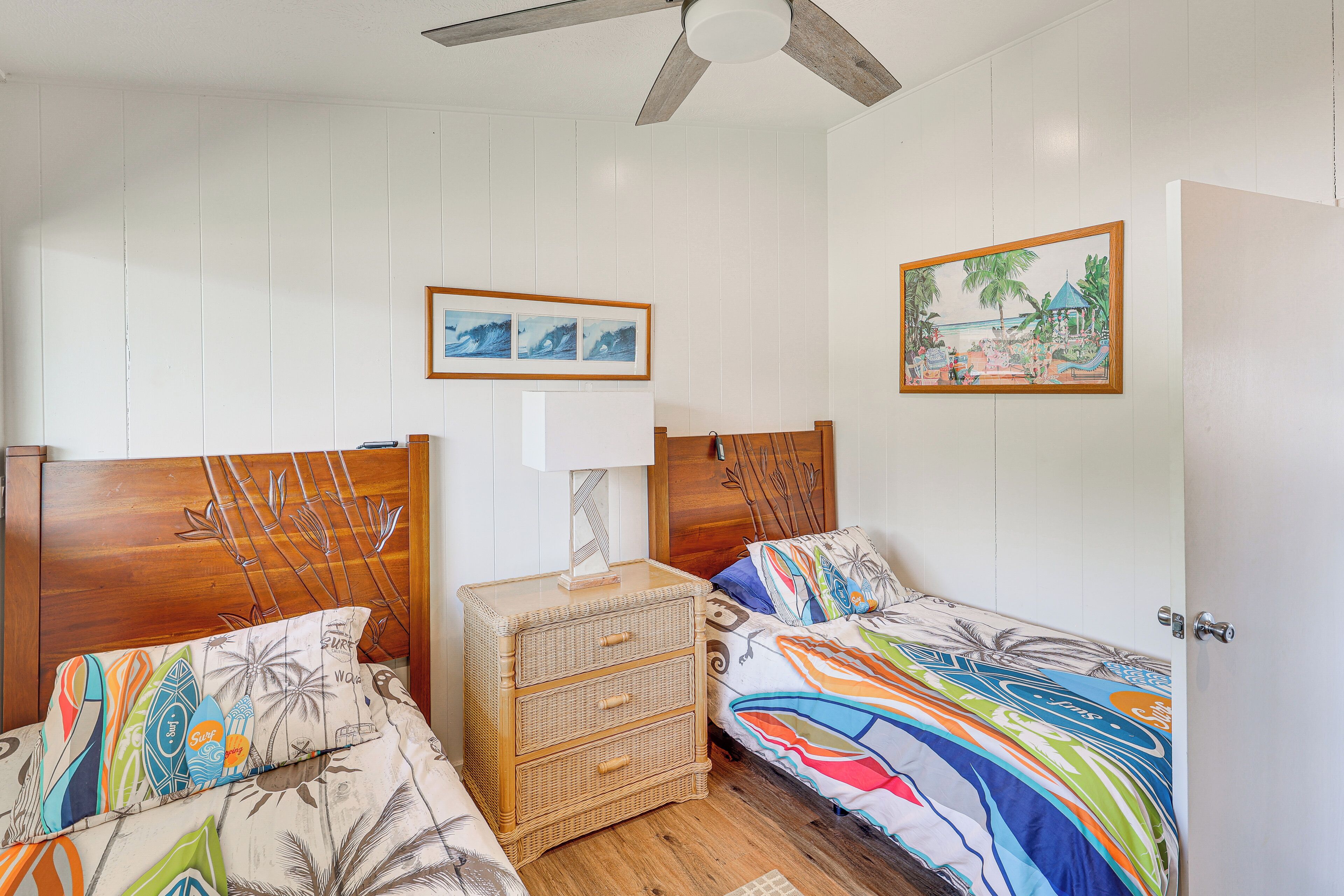 Une chambre avec deux lits simples, une table de chevet en bois avec des tiroirs, un ventilateur de plafond et un tableau accroché au mur.