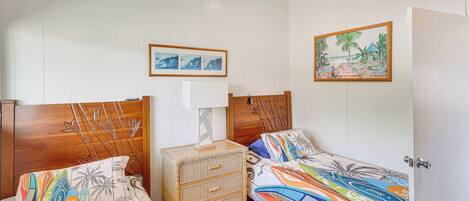 Une chambre avec deux lits simples, une table de chevet en bois avec des tiroirs, un ventilateur de plafond et un tableau accroché au mur.