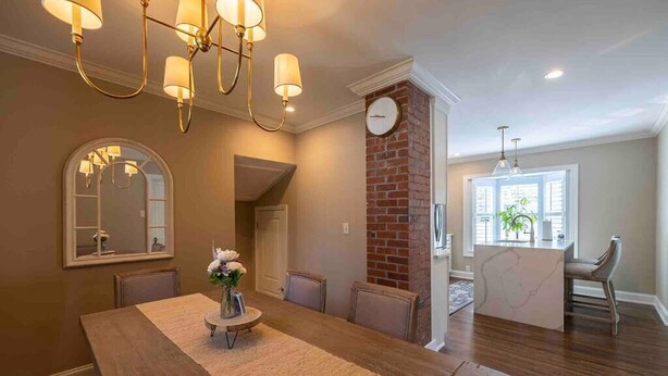 A dining area with a wooden table, chairs, a vase with flowers, a chandelier, a clock, and a brick wall.