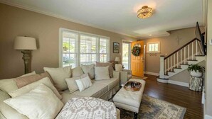 A living room with a beige sofa, a patterned throw, and a wooden coffee table. There is a staircase with a railing leading to a door with a wreath.