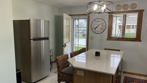 A kitchen with a stainless steel refrigerator, a wooden island with a marble countertop, wicker chairs, a clock, and a window with blinds. - Lovely Riverfront views of river. Walking distance to beach.  🚴👙🐠 (Two Rivers)