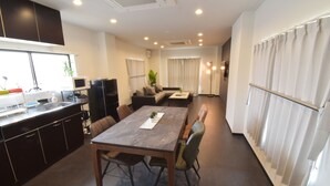 A modern kitchen with dark cabinetry, a dining area with a wooden table and chairs, and a living room with a sofa and curtains. - OguHouse Beppu (Beppu)