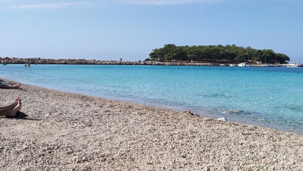 A pebbled beach with clear blue water, a small island with trees, and a rocky breakwater in the distance. - Stunning 2-Bed House in Jezera with Sea views (Jezera)