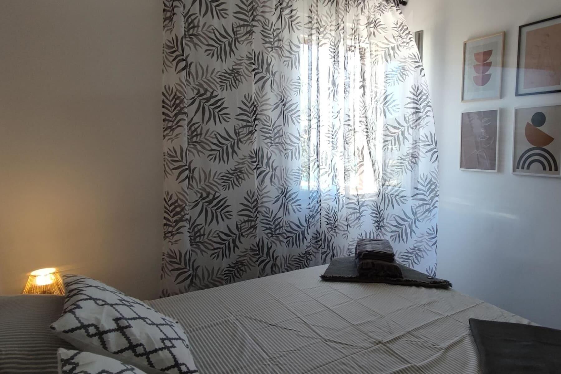 A bedroom with a bed, a nightstand, a lamp, a window with leaf-patterned curtains, and wall art.