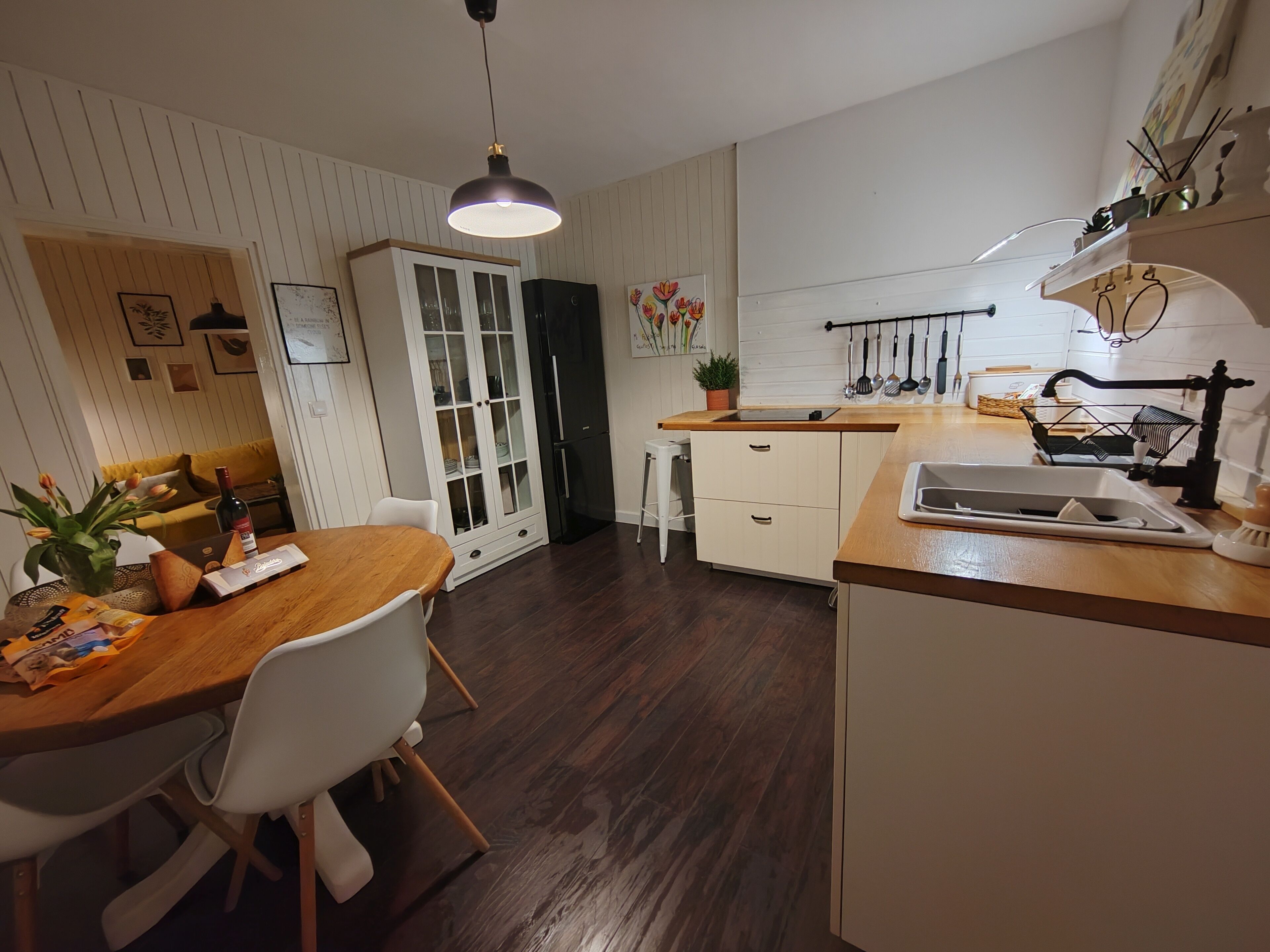 A modern kitchen with wooden countertops, a dining area with a wooden table and white chairs, a white cabinet with glass doors, and a hanging light fixture.