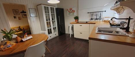A modern kitchen with wooden countertops, a dining area with a wooden table and white chairs, a white cabinet with glass doors, and a hanging light fixture.