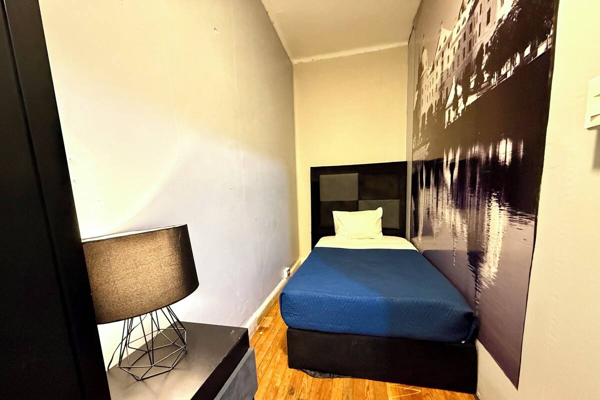 A modern hotel room with a bed, a nightstand, a lamp, and a large poster on the wall.