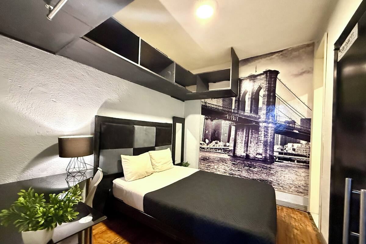 A modern bedroom with a bed, a desk, a lamp, and a large cityscape mural on the wall.