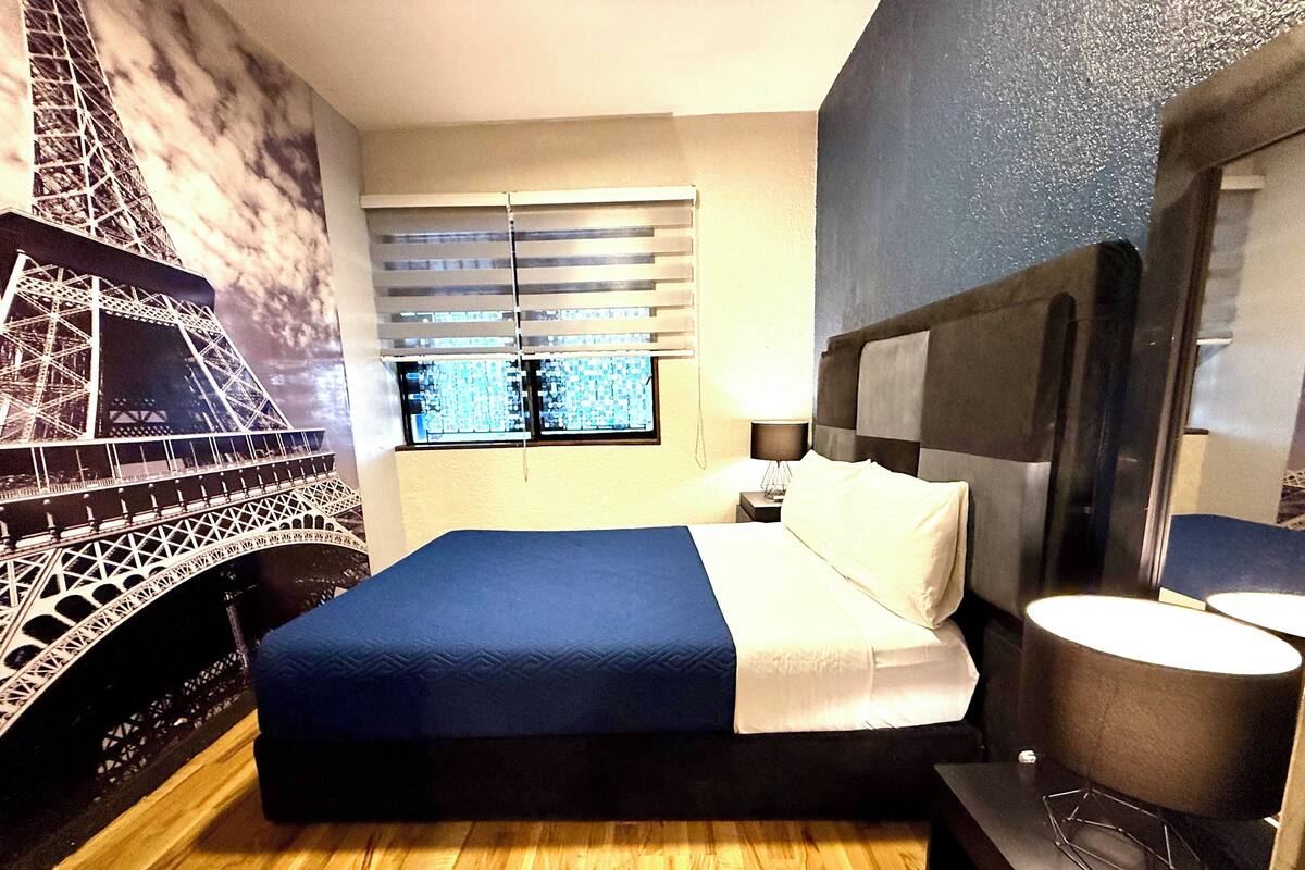 A hotel room with a bed, a nightstand, a lamp, a window with blinds, and a wall mural of the Eiffel Tower.