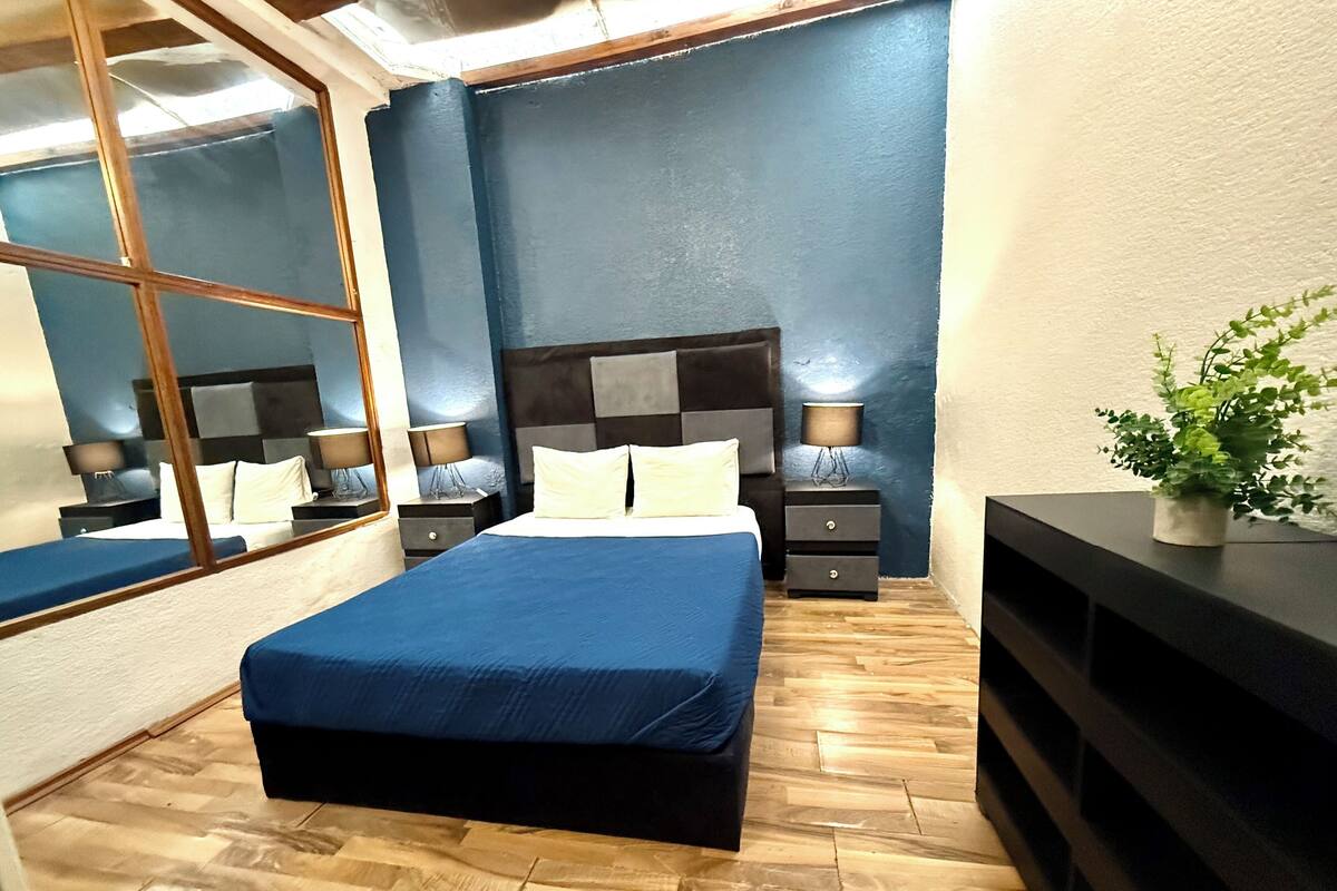 A modern bedroom with a large bed, a wooden floor, a dark blue accent wall, and a large mirror.