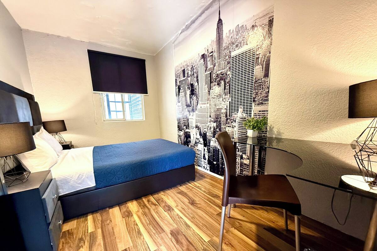 A compact hotel room with a bed, a desk, a chair, a window, and a cityscape mural on the wall.