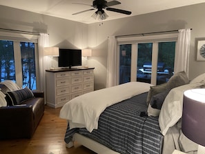 Un dormitorio con una cama, un televisor sobre un aparador, una silla y una ventana con cortinas.