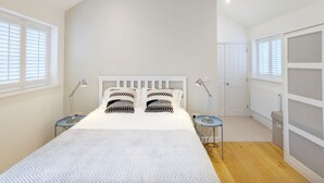 Ein Schlafzimmer mit schrägen Wänden, einem Bett mit weißem Kopfteil, zwei Nachttischlampen und einem Fenster mit Jalousien.