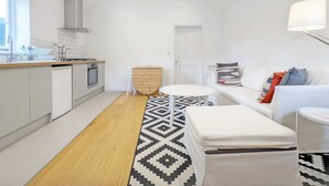 Eine moderne Küche mit weißen Schränken, einer Holz Arbeitsplatte und einem eingebauten Ofen. Ein Essbereich mit weißem Tisch und Stühlen lädt zum Verweilen ein, und ein gemustertes Teppich liegt auf dem Boden. Gegenüber der Wand steht ein weißes Sofa mit bunten Kissen, daneben ein Nachttisch mit einer weißen Lampe.
