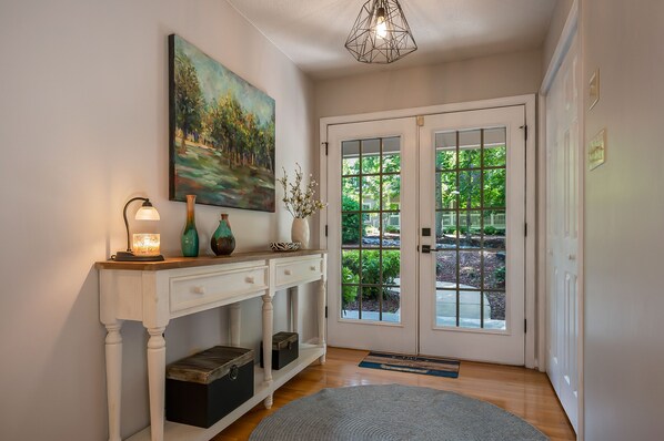 Un couloir avec une console blanche, un tableau, une porte à panneaux de verre et une vue sur la verdure à l’extérieur.