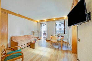 A living room with a sofa, a dining table with chairs, a TV mounted on the wall, and a window with a view of buildings. - Monterosa Cozy Apartment 200m From Ski, Macugnaga, Italy (Macugnaga)