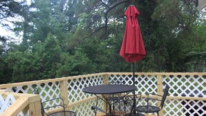 Una terraza de madera con barandilla de celosía blanca, una mesa redonda con cuatro sillas y un paraguas rojo.
