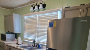 Una cocina con un refrigerador de acero inoxidable, lavavajillas, microondas y un lavamanos con grifo. Hay armarios y una ventana con persianas.