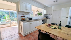 A modern kitchen with white cabinets, a wooden dining table set with food and wine, and a view of the outdoors.