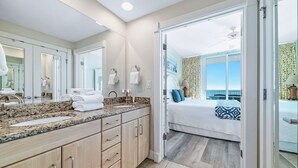 Un baño de hotel con un espejo grande, un lavabo y tocador. Se observa una cama con vista al océano y un balcón con mesa y sillas.