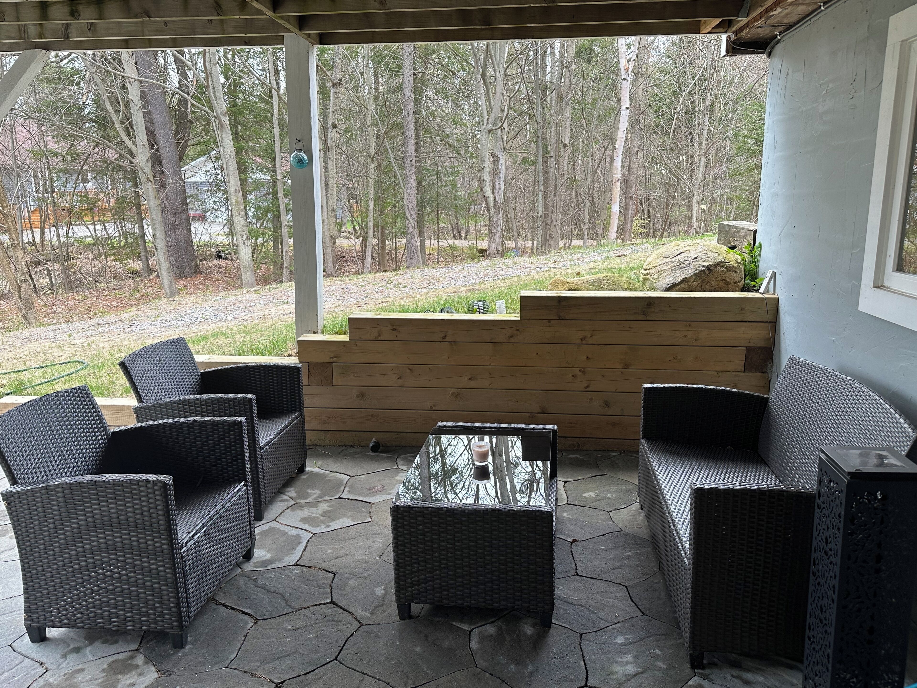 A covered patio area with wicker furniture and a stone floor, overlooking a wooded area.